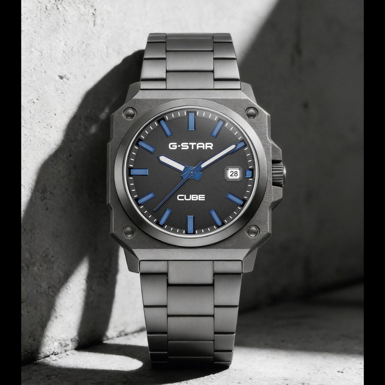A G-Star Cube watch with a dark gray metallic case and link bracelet, featuring a blue dial with blue markers and hands, resting against a textured concrete wall in dramatic shadow.