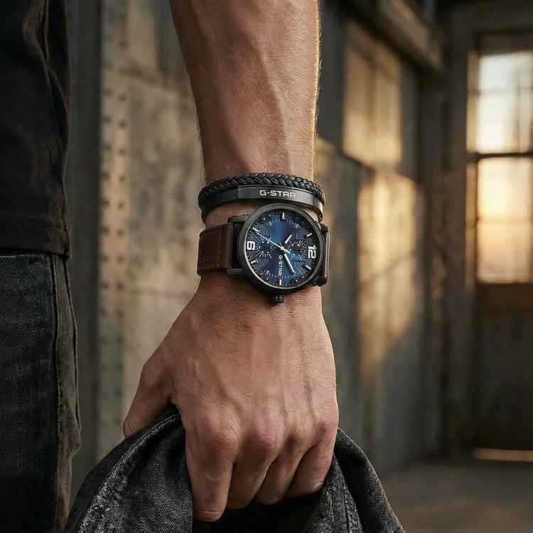A close-up lifestyle shot of a man's wrist wearing a Stilo brand watch with a dark blue dial and brown leather strap, styled with a black braided bracelet. He is holding a dark denim jacket in a dimly lit, rustic industrial setting.