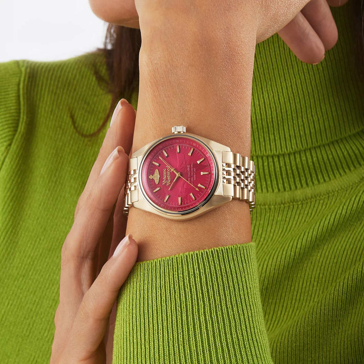 A close-up of a woman's wrist wearing a gold-tone watch with a vibrant pink dial and matching pink bezel. The watch has gold-tone hour markers and hands.