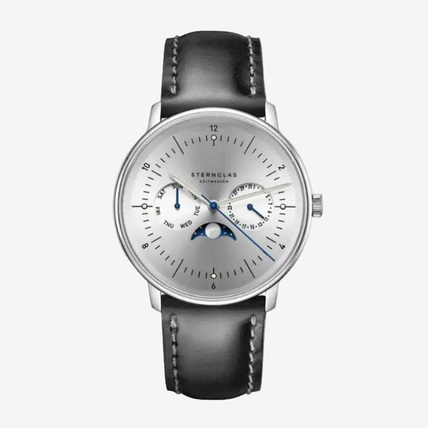 Sternglas Astronom watch with a silver dial, blue hands, moon phase indicator, and a black leather strap with white stitching, shown on a light gray background.