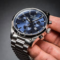 A close-up of a silver-toned stainless steel watch with a blue dial and matching blue sub-dials, held in a person's hand against a dark gray gradient background.