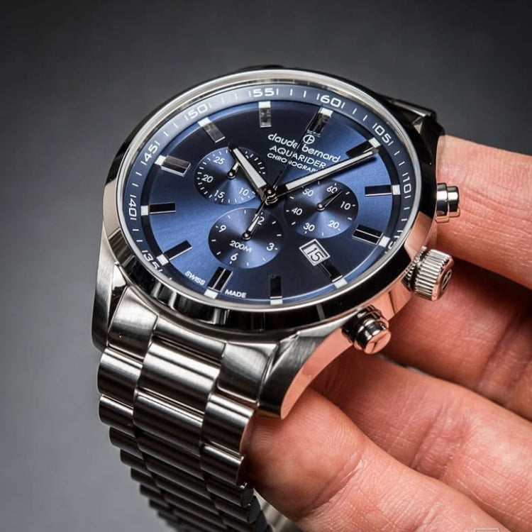 A close-up of a silver-toned stainless steel watch with a blue dial and matching blue sub-dials, held in a person's hand against a dark gray gradient background.