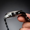 Close-up of a silver stainless steel watch case held by a hand, showing the side profile with three screw details and a black-and-silver patterned strap against a dark gray background.