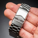A close-up of a stainless steel watch band clasp held in a hand, showing the brushed and polished metal surfaces against a dark gray background.