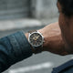 A close-up lifestyle shot of a man wearing a silver-toned chronograph watch with a black dial and brown leather strap, shown on his wrist against a blurred street background.