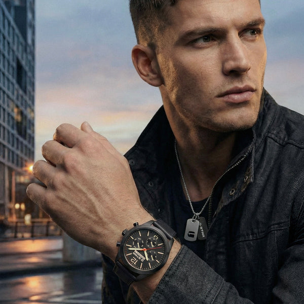 A man in a dark denim jacket wearing a black chronograph watch with a black strap, photographed outdoors against a city street at dusk.