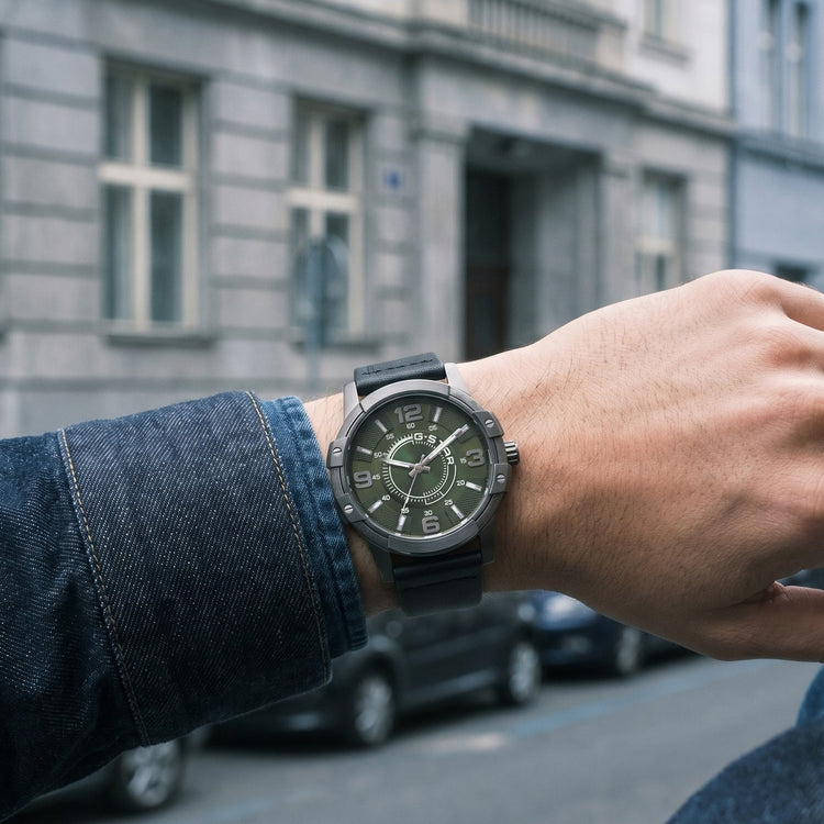 A close-up of a person's wrist wearing a black military-style watch with a dark dial and black strap, set against a blurred street background with buildings and cars.