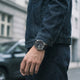 A close-up lifestyle shot of a person wearing a dark navy denim jacket and matching dark trousers, with a wristwatch on the wrist. The watch has a dark dial and a silver-toned metal case, set against a blurred street background with a gray car and building.