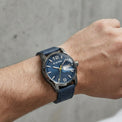 A wristwatch with a textured blue dial and a dark blue strap, worn on a person's wrist against a gray concrete wall background.