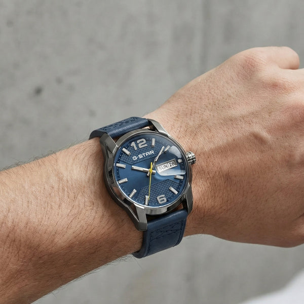 A wristwatch with a textured blue dial and a dark blue strap, worn on a person's wrist against a gray concrete wall background.