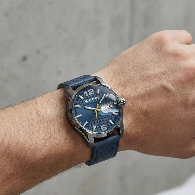 A wristwatch with a textured blue dial and a dark blue strap, worn on a person's wrist against a gray concrete wall background.