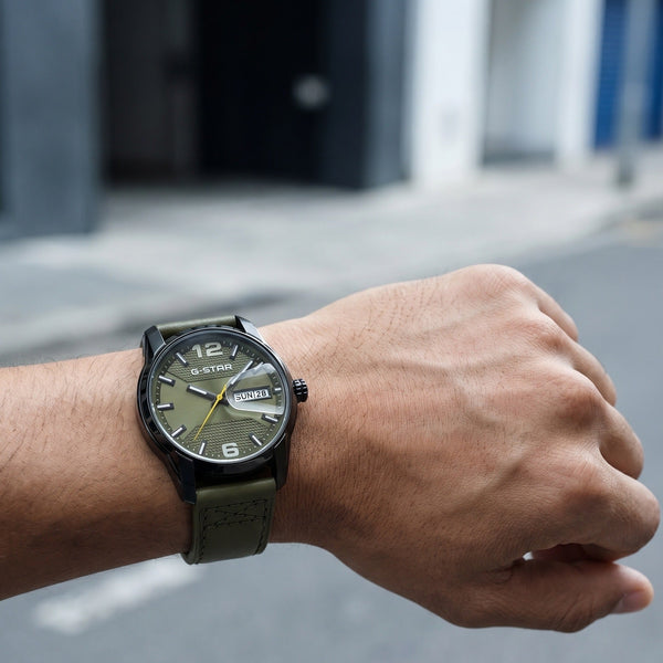 A wristwatch with a dark olive green strap and a black dial, worn on a person's wrist against a blurred outdoor street background.