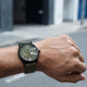 A wristwatch with a dark olive green strap and a black dial, worn on a person's wrist against a blurred outdoor street background.