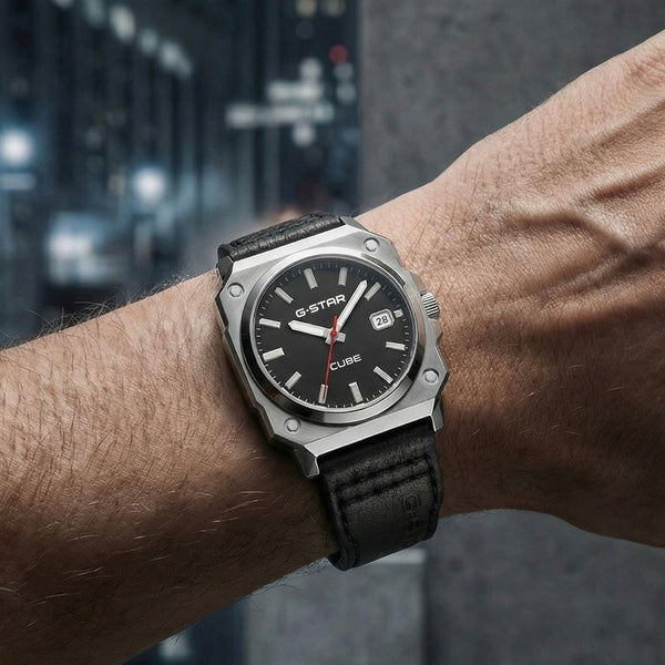 A wristwatch with a black dial, silver-toned case, and black strap worn on a person's wrist, photographed against a blurred urban background.