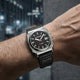 A wristwatch with a black dial, silver-toned case, and black strap worn on a person's wrist, photographed against a blurred urban background.