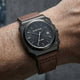 A wristwatch with a dark gray case, black dial, and brown leather strap, shown worn on a person's wrist against a dark gray background.
