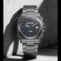A G-Star Cube watch with a dark gray metallic case and link bracelet, featuring a blue dial with blue markers and hands, resting against a textured concrete wall in dramatic shadow.