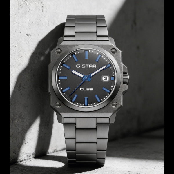 A G-Star Cube watch with a dark gray metallic case and link bracelet, featuring a blue dial with blue markers and hands, resting against a textured concrete wall in dramatic shadow.