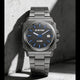 A G-Star Cube watch with a dark gray metallic case and link bracelet, featuring a blue dial with blue markers and hands, resting against a textured concrete wall in dramatic shadow.