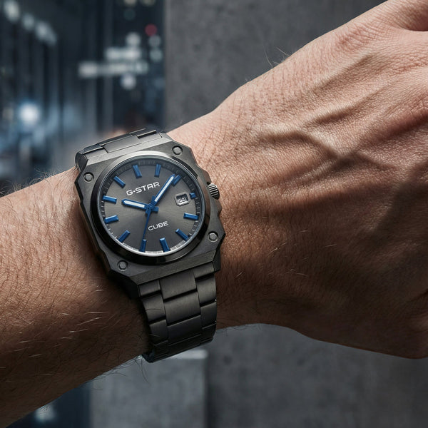 A wrist-worn Oyster Quartz watch with a dark gray metal case and bracelet, featuring a blue dial and blue hands, shown on a person's wrist against a blurred dark background.