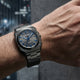 A wrist-worn Oyster Quartz watch with a dark gray metal case and bracelet, featuring a blue dial and blue hands, shown on a person's wrist against a blurred dark background.