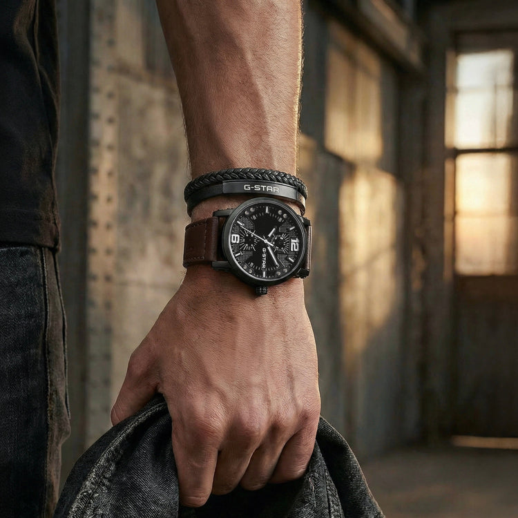 A close-up lifestyle shot of a man's hand wearing a U-Strönd black analog watch with a brown leather strap and black braided bracelet, holding a dark denim jacket in a dimly lit, rustic industrial setting.