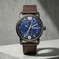 A wristwatch with a dark blue textured dial, silver-tone hands and markers, and a brown leather strap, displayed on a gray concrete block against a gray wall.