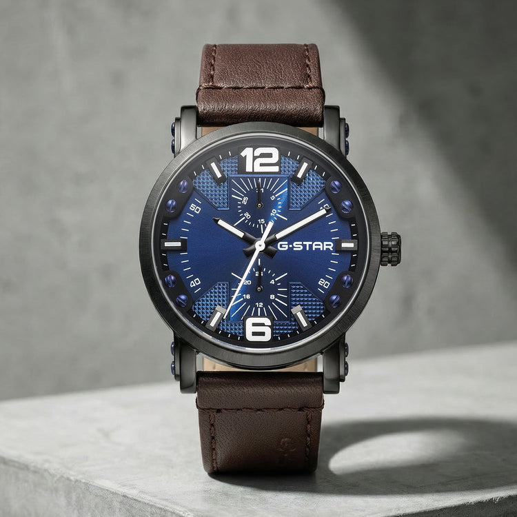 A wristwatch with a dark blue textured dial, silver-tone hands and markers, and a brown leather strap, displayed on a gray concrete block against a gray wall.