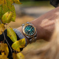 Gold and green watch on a wrist with autumn leaves in the background