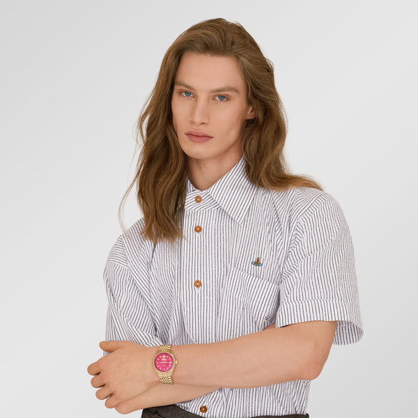 A model with long light brown hair is wearing a short-sleeve button-down shirt with thin blue and white vertical stripes, brown buttons, and a small embroidered logo on the chest. He is also wearing dark gray trousers and a gold watch with a pink dial.