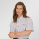 A model with long light brown hair is wearing a short-sleeve button-down shirt with thin blue and white vertical stripes, brown buttons, and a small embroidered logo on the chest. He is also wearing dark gray trousers and a gold watch with a pink dial.