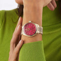 A close-up of a woman's wrist wearing a gold-tone watch with a vibrant pink dial and matching pink bezel. The watch has gold-tone hour markers and hands.