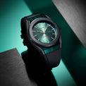 A sleek wristwatch with a dark gray case, a green dial, and a green translucent strap, resting on a dark teal and black background with brushed metal panels.