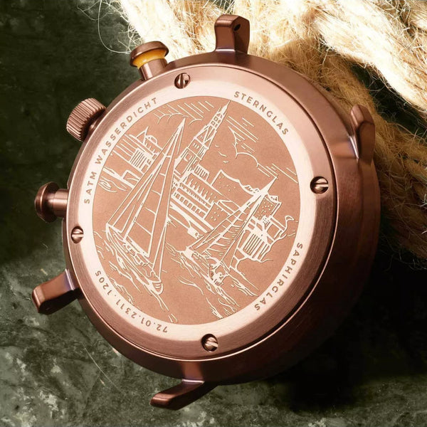 A close-up of a bronze-toned watch back casing with engraved nautical ship artwork and text, resting against a dark textured surface with beige rope in the background.