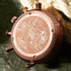 A close-up of a bronze-toned watch back casing with engraved nautical ship artwork and text, resting against a dark textured surface with beige rope in the background.