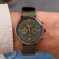 A close-up of a wrist wearing a military-style chronograph watch with an olive green dial, matching green fabric strap, and gold-tone hands.