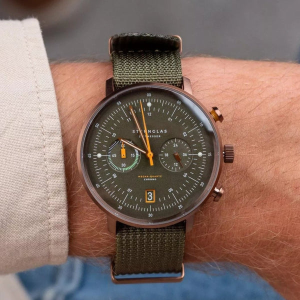 A close-up of a wrist wearing a military-style chronograph watch with an olive green dial, matching green fabric strap, and gold-tone hands.