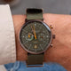 A close-up of a wrist wearing a military-style chronograph watch with an olive green dial, matching green fabric strap, and gold-tone hands.