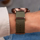 A close-up of a watch with a textured olive green fabric strap and a rose gold case, worn on a person's wrist.