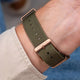An olive green woven canvas watch strap with a rose gold buckle, shown worn on a person's wrist.