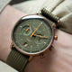 A close-up of a wrist wearing a Ströndahl watch with an olive green dial, rose gold-toned case, and olive green fabric strap, set against a neutral beige background.